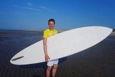 Portrait of man with surfboard standing at beach against blue sky