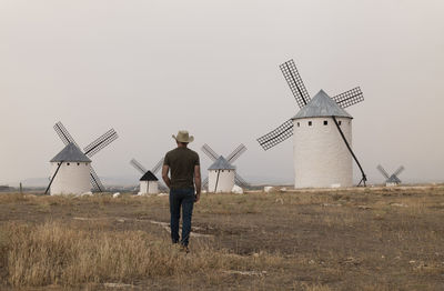 Rear view of man with cowboy hat standing on field with spanish white windmills against sky