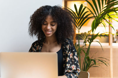 Young cheerful ethnic female with afro hairstyle browsing internet on portable computer against palm plant
