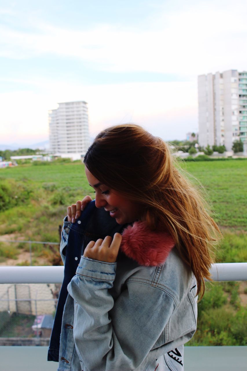 city, leisure activity, railing, lifestyles, architecture, connection, built structure, casual clothing, young adult, sky, young women, building exterior, holding, long hair, modern, focus on foreground, cloud - sky, person, tourism, skyscraper, tall - high, day