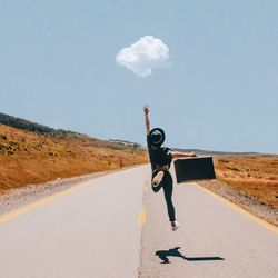 Full length rear view of woman jumping while holding luggage on road against sky