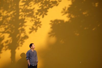 Mid adult man looking away while standing by yellow wall