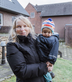 Portrait of smiling woman carrying girl standing against building during winter