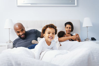 Parents playing with children while relaxing on bed against wall at home