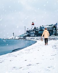 Rear view of man walking on shore against buildings during snowfall