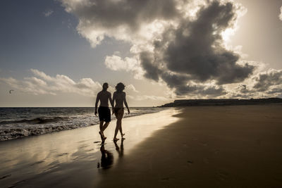 Rear view of man and woman holding hands while walking at the beach during sunset