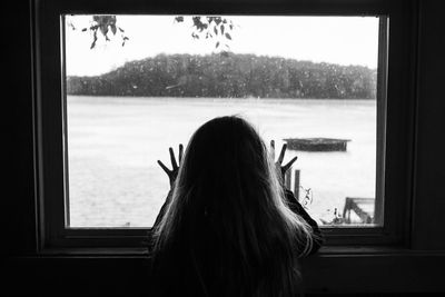 Rear view silhouette of young girl looking through window at a lake