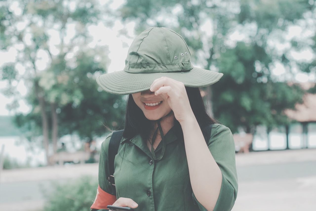 hat, one person, clothing, real people, focus on foreground, smiling, front view, leisure activity, lifestyles, young adult, casual clothing, happiness, waist up, portrait, day, adult, standing, plant, outdoors, hairstyle