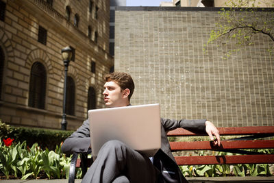 Businessman with laptop computer looking away while sitting on bench against buildings