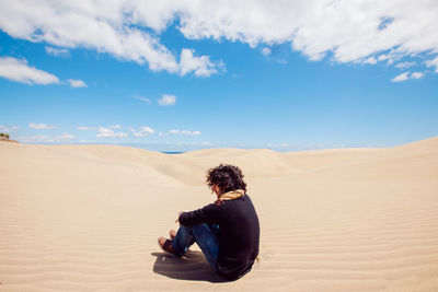 Man sitting on sand in desert against sky