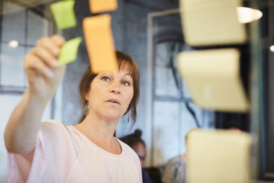 Mature businesswoman sticking sticky note on glass in creative office