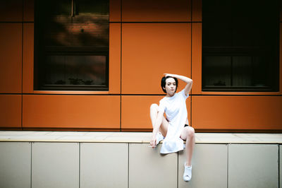 Full length of young woman sitting on walkway against orange building