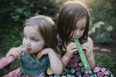 Sisters looking away while sucking plastic tube popsicles at yard