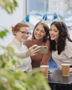 Smiling woman wearing birthday cap taking selfie with friends at cafe