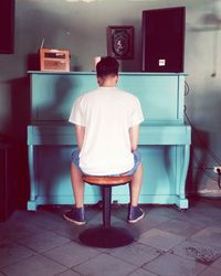 Rear view full length of man sitting by turquoise grand piano at home