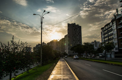 Road by buildings in city against sky during sunset