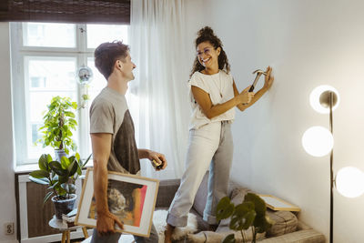 Happy girlfriend with hammer looking at boyfriend holding frame at home