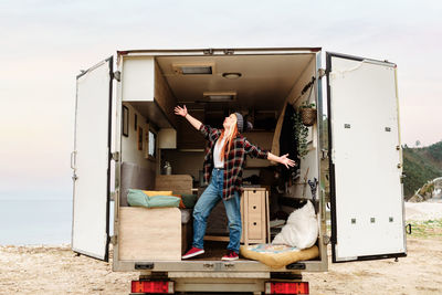 Side view of dreamy traveling female hipster standing inside of truck parked near sea in mountainous area and enjoying vacation with outstretched arms