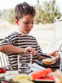 Focused adorable child smearing butter on slice of bread while having breakfast at table in courtyard in summer