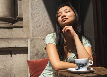 Dreamy asian female with closed eyes sitting at table with cup of coffee on terrace of outdoor cafe on sunny summer day and enjoying weekend