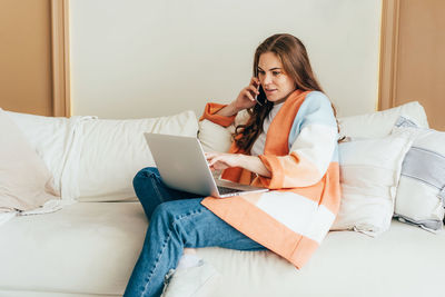 Young business woman in a cardigan sitting on the couch works on a laptop and speaks on the phone.