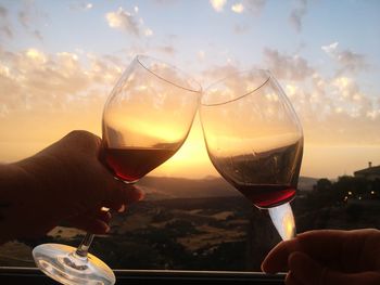 Close-up of hand holding wineglasses against sky during sunset