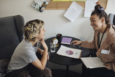 Smiling non-binary psychologist looking at teenage student discussing while sitting in school office