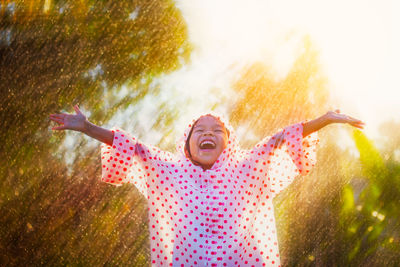 Happy girl wearing raincoat with arms outstretched standing in park during rainfall