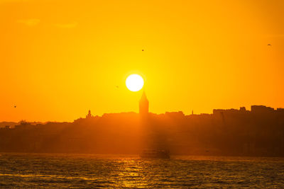 Silhouette buildings by river against orange sky