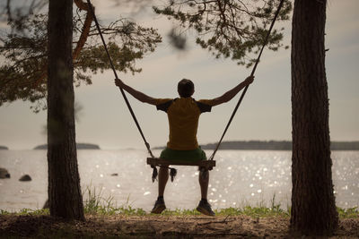 The boy is swinging on a swing on the shore of the lake. the swing is suspended from trees.