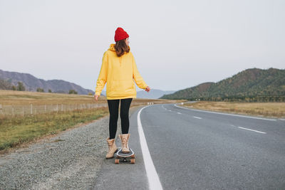 Young woman in yellow hoodie and red hat on skateboard on road against beautiful mountain 