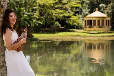 Smiling young woman playing ukelele while standing by lake