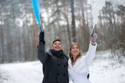 Smiling wedding couple holding distress flares while standing at forest during winter