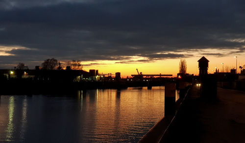 Silhouette buildings by river against sky at sunset