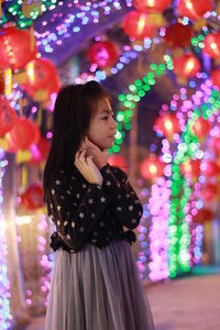 Girl looking away while standing against illuminated lights