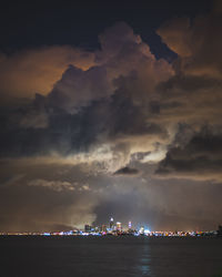 Illuminated city by sea against sky at sunset