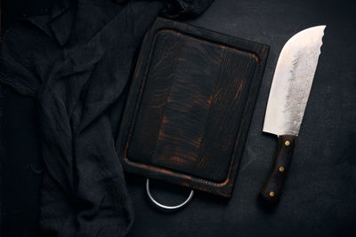 Rectangular empty wooden cutting board and kitchen knife on black table with gauze napkin, top view