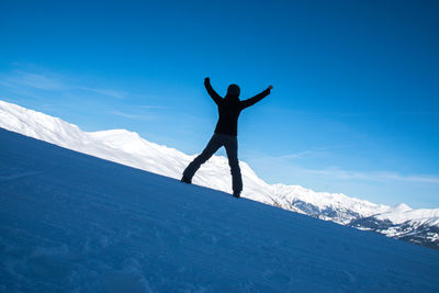 Full length of excited woman with arms raised standing against snowcapped mountain and blue sky