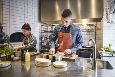 Young male chef preparing food by female colleagues at counter in restaurant kitchen