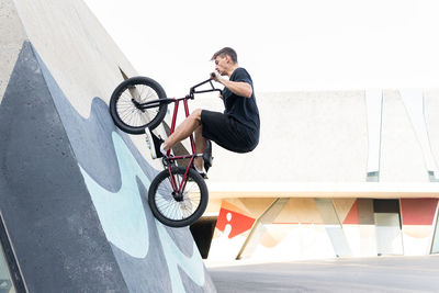 Side view of young male in casual clothes riding concrete wall on bmx bicycle while spending time in skate park in weekend