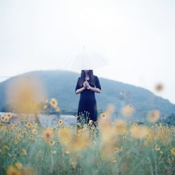 Woman holding umbrella while standing amidst yellow flowering plants on field by mountain against sky