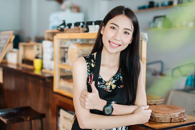 Portrait of smiling young woman with arms crossed gesturing thumbs up at home