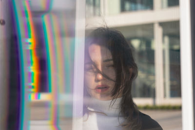 Portrait of young woman standing against building in city seen through window