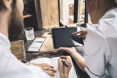 Midsection of chef with digital tablet while coworker writing at table in restaurant