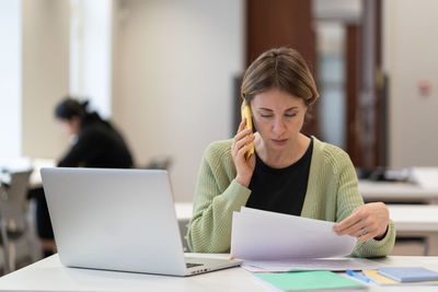 Middle-aged woman busy with documents talking on cellphone working in campus open space. education.