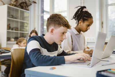 Focused schoolboy using laptop by female friend at desk in classroom