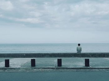 Rear view of man sitting on retaining wall at promenade against cloudy sky