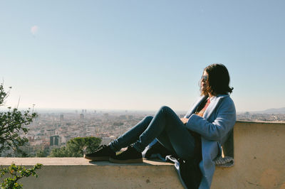 Woman sitting on retaining wall against clear sky