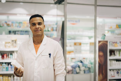 Portrait of confident owner wearing lab coat standing with medicine in pharmacy store