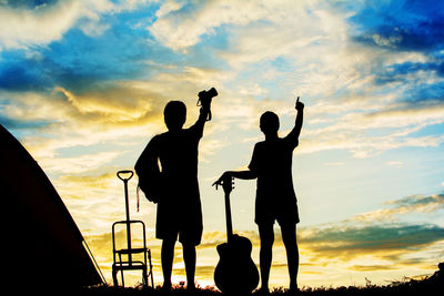 Silhouette couple with guitar and camera gesturing towards cloudy sky